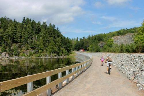 Two people biking along a scenic path beside a calm lake, surrounded by trees and rocky terrain under a blue sky.