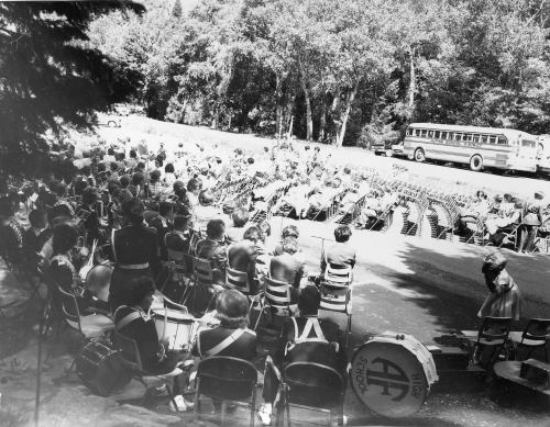 A large crowd seated in rows, watching a performance, with a bus parked nearby and trees in the background.
