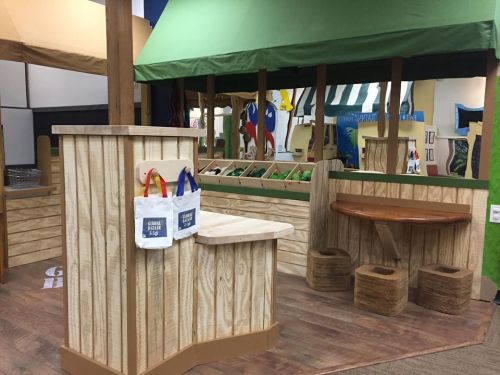 A wooden play area featuring a market stall with colorful bags and a seating area, designed for children's imaginative play.