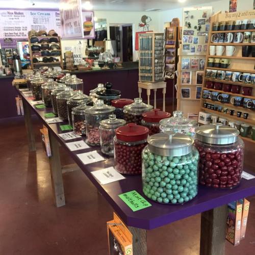 A colorful display of jars filled with candies on a purple table in a shop, with various products and decorations in the background.