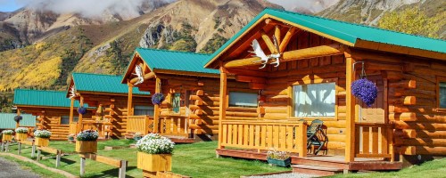 Three rustic log cabins with green roofs, surrounded by colorful autumn foliage and mountains in the background.