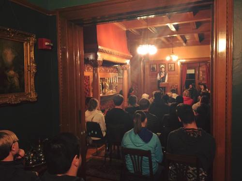 A cozy room with a speaker at the front, addressing an audience seated in rows, surrounded by vintage decor.