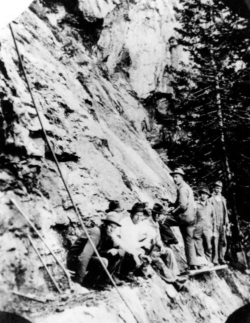 Black and white photo of a group of miners sitting and standing near a rocky cliff in a forested area.