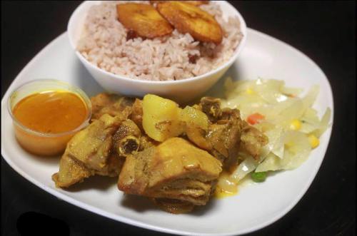 A plate of rice with chicken, plantains, and a side of mixed vegetables, accompanied by a small container of sauce.