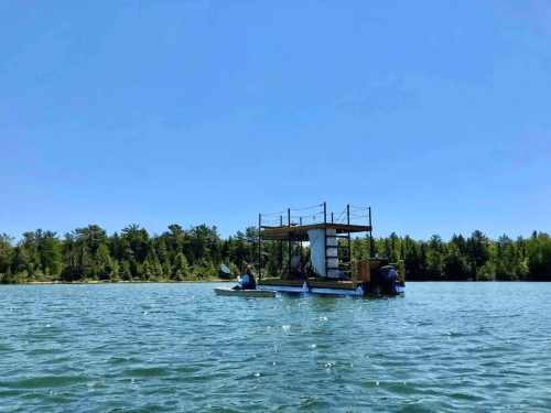 A small boat with people paddling near a floating structure on a calm lake, surrounded by trees under a clear blue sky.