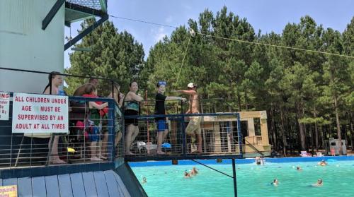 A group of children and adults at a swimming area, with a diving platform and trees in the background.