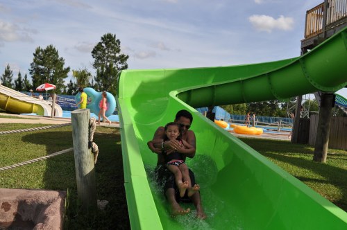 A child and adult slide down a green water slide, splashing into the water at a water park on a sunny day.