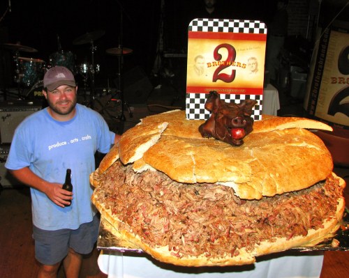 A man stands next to an enormous pulled pork sandwich topped with a small figurine, holding a drink in his hand.