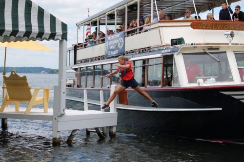 A person in a red shirt leaps from a boat to a dock on a lake, with passengers watching from the boat.
