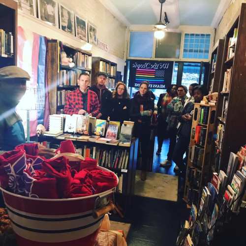 A cozy bookstore interior with a diverse group of people gathered, listening to a speaker near a display of books.