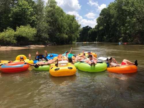 A group of people relaxing on colorful inner tubes in a river, surrounded by trees and a sunny sky.