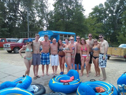 A group of young adults in swimwear poses by a river with inflatable tubes, surrounded by trees and parked vehicles.
