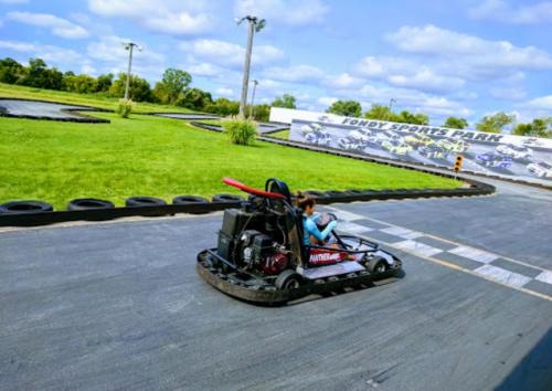 A go-kart racing on a track at Fondy Sports Park, with green grass and a cloudy sky in the background.