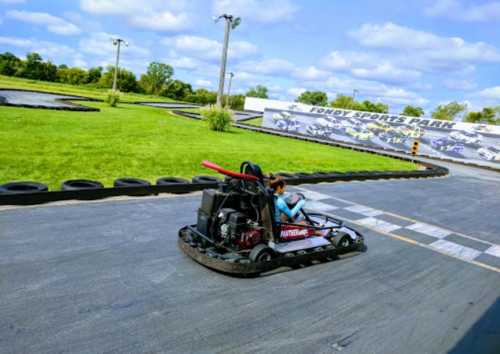 A go-kart racing on a track at Fondy Sports Park, with green grass and a cloudy sky in the background.