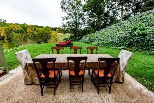 A dining table with chairs set outdoors, surrounded by greenery and a view of a lawn and distant trees.