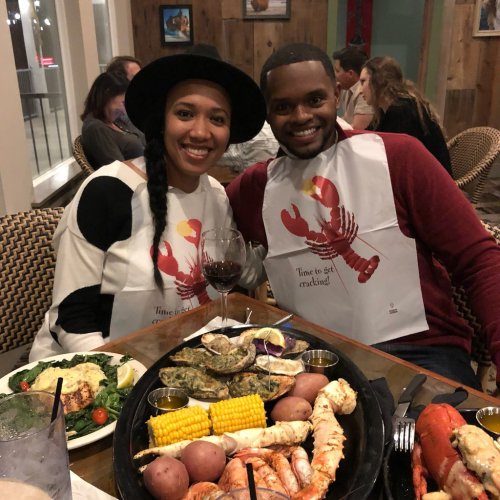 A couple smiles at the camera, wearing bibs, with a seafood feast on the table and a glass of wine nearby.