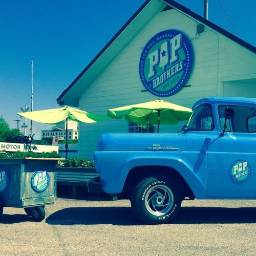 A blue vintage truck parked outside a building with "Pop Brothers" signage and green umbrellas.