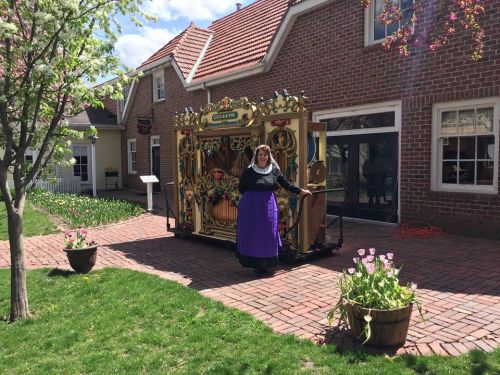 A woman in traditional attire stands beside a decorative street organ in a garden with blooming flowers and brick pathways.