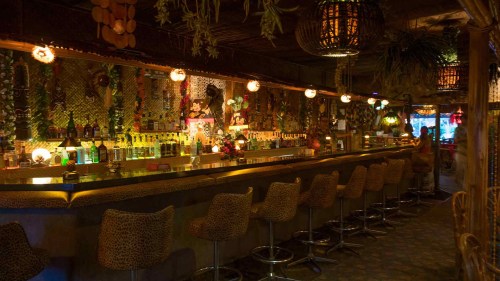 A dimly lit tiki bar with a long counter, colorful drinks, and tropical decor, featuring hanging lights and plants.