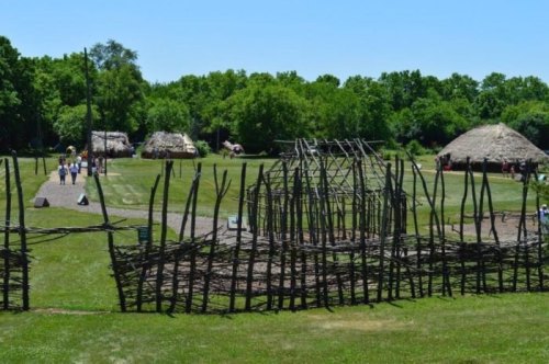 A historical village with thatched huts and wooden structures, surrounded by green trees and a clear blue sky.