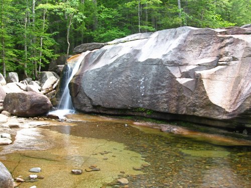 A serene waterfall cascades over a large rock into a clear pool, surrounded by lush green trees.