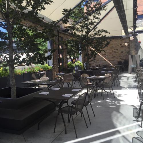 Outdoor restaurant patio with tables, chairs, and trees, featuring a modern design and shaded seating area.