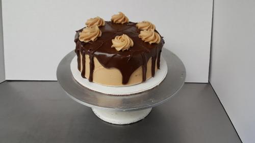 A round chocolate cake with a glossy ganache topping and decorative swirls of frosting on top, displayed on a silver stand.