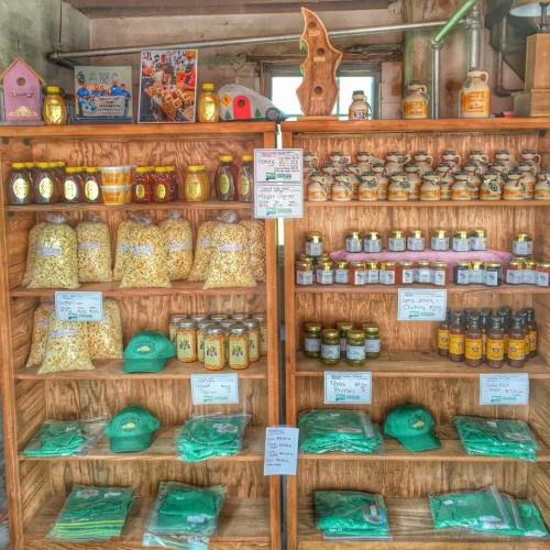 A wooden shelf displays jars of honey, bags of popcorn, and green hats, all labeled with prices.