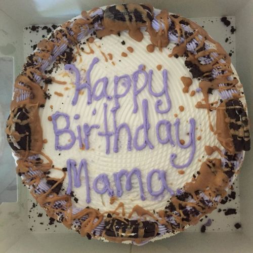 A round birthday cake with white frosting, purple writing that says "Happy Birthday Mama," and chocolate toppings.