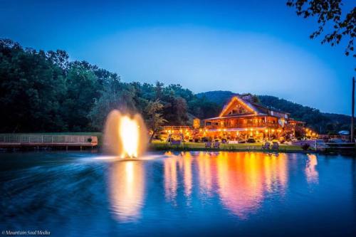 A serene lakeside scene at dusk, featuring a lit lodge and a fountain reflecting colorful lights on the water.