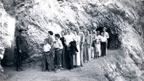 A line of people, including children and adults, waiting near a rocky cave entrance, with a soldier in uniform.