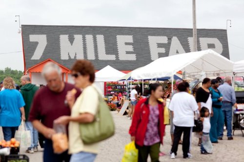 A bustling outdoor market scene with people shopping and colorful tents, featuring a large sign that reads "7 Mile Fair."