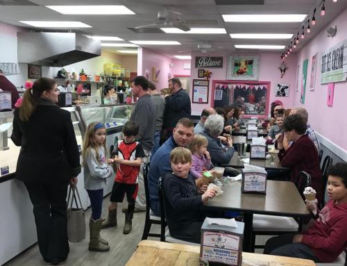 A busy ice cream shop with families and children enjoying ice cream at tables and a line at the counter.