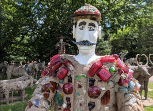 A large, colorful statue made of stone and glass, featuring a man with a mustache and a cap, surrounded by other sculptures.