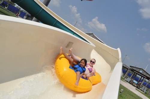 Two people in yellow inner tubes slide down a water slide on a sunny day, splashing water around them.