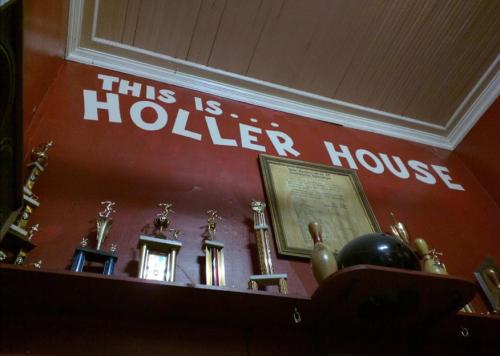A red wall with the text "THIS IS... HOLLER HOUSE" and various trophies displayed on a shelf below.