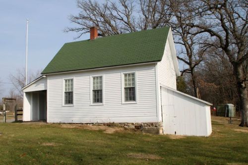 A small white house with a green roof, featuring multiple windows and a side extension, set in a grassy area.