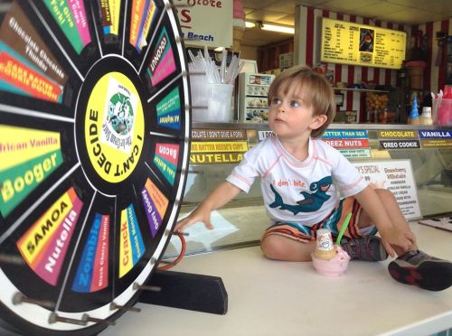 A young boy sits on an ice cream counter, reaching for a colorful spinning wheel while holding an ice cream cone.