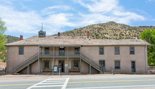 Historic building with two stories, stairs on both sides, surrounded by mountains and trees under a blue sky.