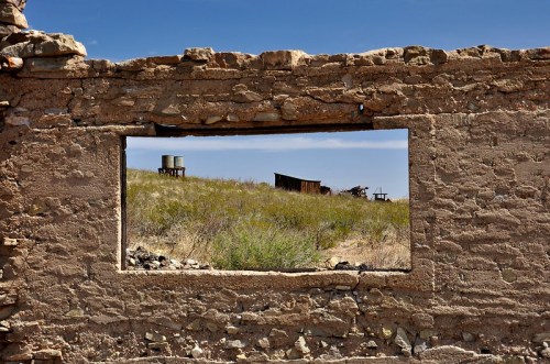 View through a weathered wall, framing a grassy landscape with an old water tank and abandoned structures in the distance.