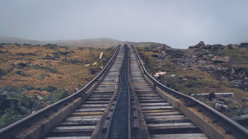 A foggy landscape with wooden train tracks stretching into the distance, surrounded by rocky terrain and sparse vegetation.