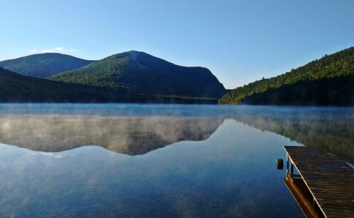 A serene lake reflects mountains under a clear blue sky, with mist rising from the water and a wooden dock in the foreground.