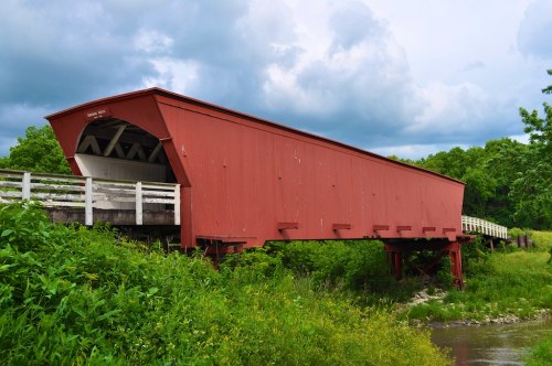 A red covered bridge stands over a creek, surrounded by lush greenery and under a cloudy sky.