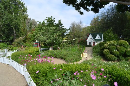 A vibrant garden with colorful flowers, lush greenery, and a charming cottage in the background under a blue sky.