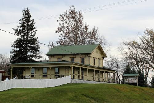 A yellow two-story house with a green roof, surrounded by trees and a white fence, on a grassy hill.