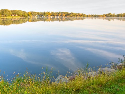 A calm lake reflects trees and a clear sky, with grassy shores in the foreground.