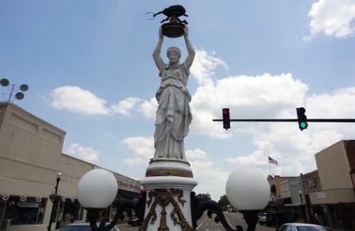 A tall white statue holding a figure atop a pedestal, with a green traffic light and buildings in the background.