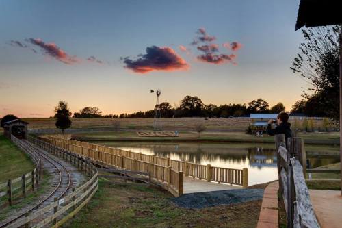 A serene landscape at sunset featuring a pond, wooden bridge, and a person taking photos near a railway track.