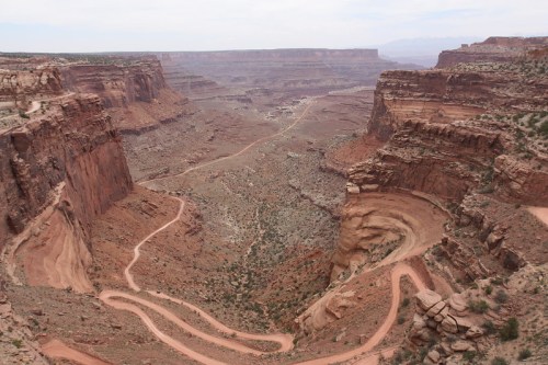 A winding dirt road cuts through a vast canyon landscape with rocky cliffs and sparse vegetation under a cloudy sky.