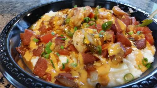 A black bowl filled with shrimp, diced tomatoes, green onions, and melted cheese over creamy grits.
